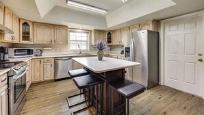Une cuisine moderne avec des meubles en bois, un îlot central doté de tabourets de bar, des appareils électroménagers en acier inoxydable et une grande fenêtre qui laisse entrer la lumière naturelle.