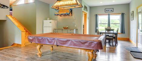 A pool table in a room with a wooden floor, a ceiling fan, and a dining area with a table and chairs.