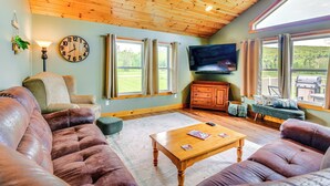 Een gezellige woonkamer met een houten plafond, een grote klok, een flatscreen televisie en uitzicht op het landschap door grote ramen.