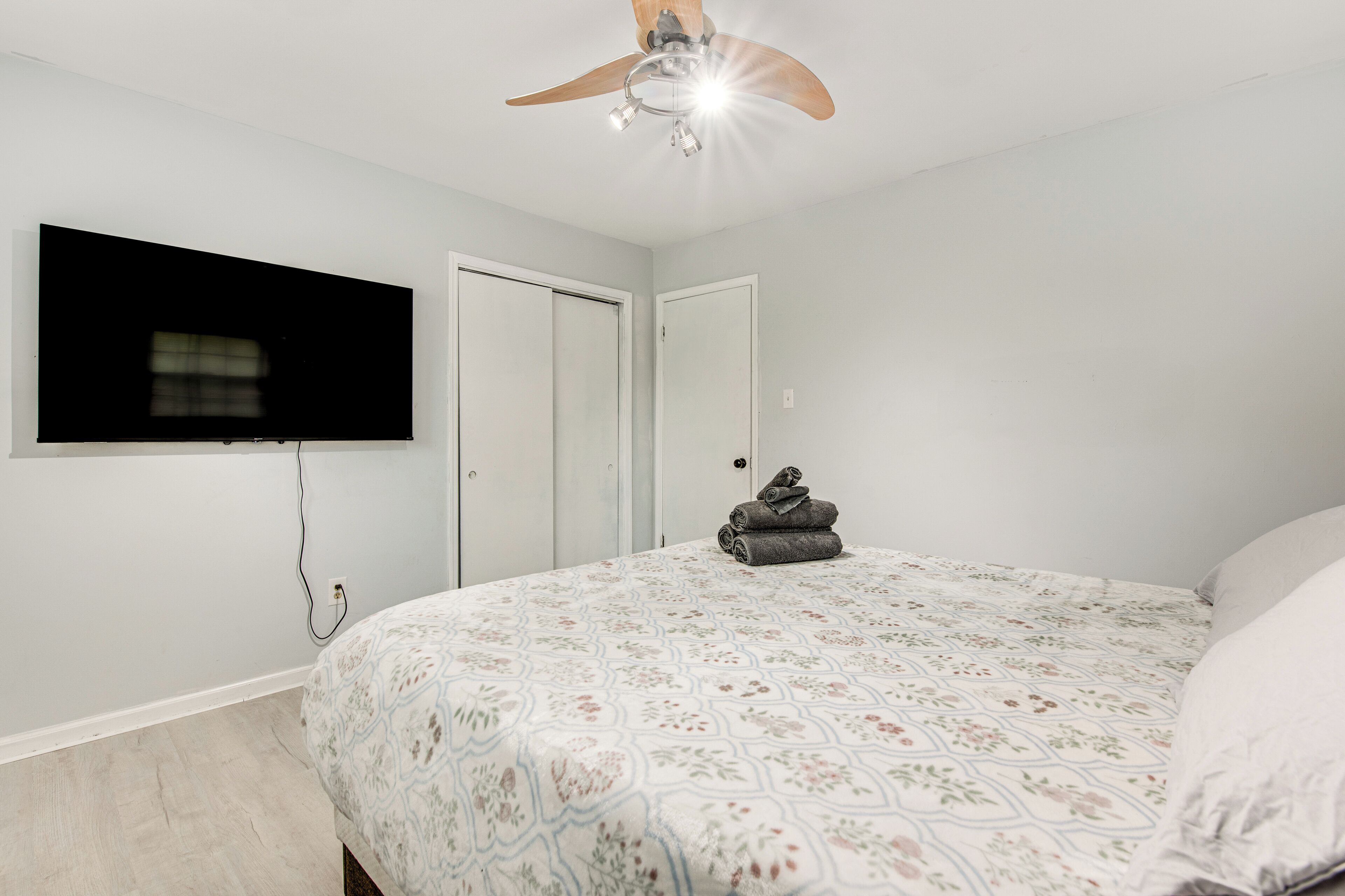 Ein Schlafzimmer mit Bett, Deckenventilator, an der Wand montiertem Fernseher und einer Tür.