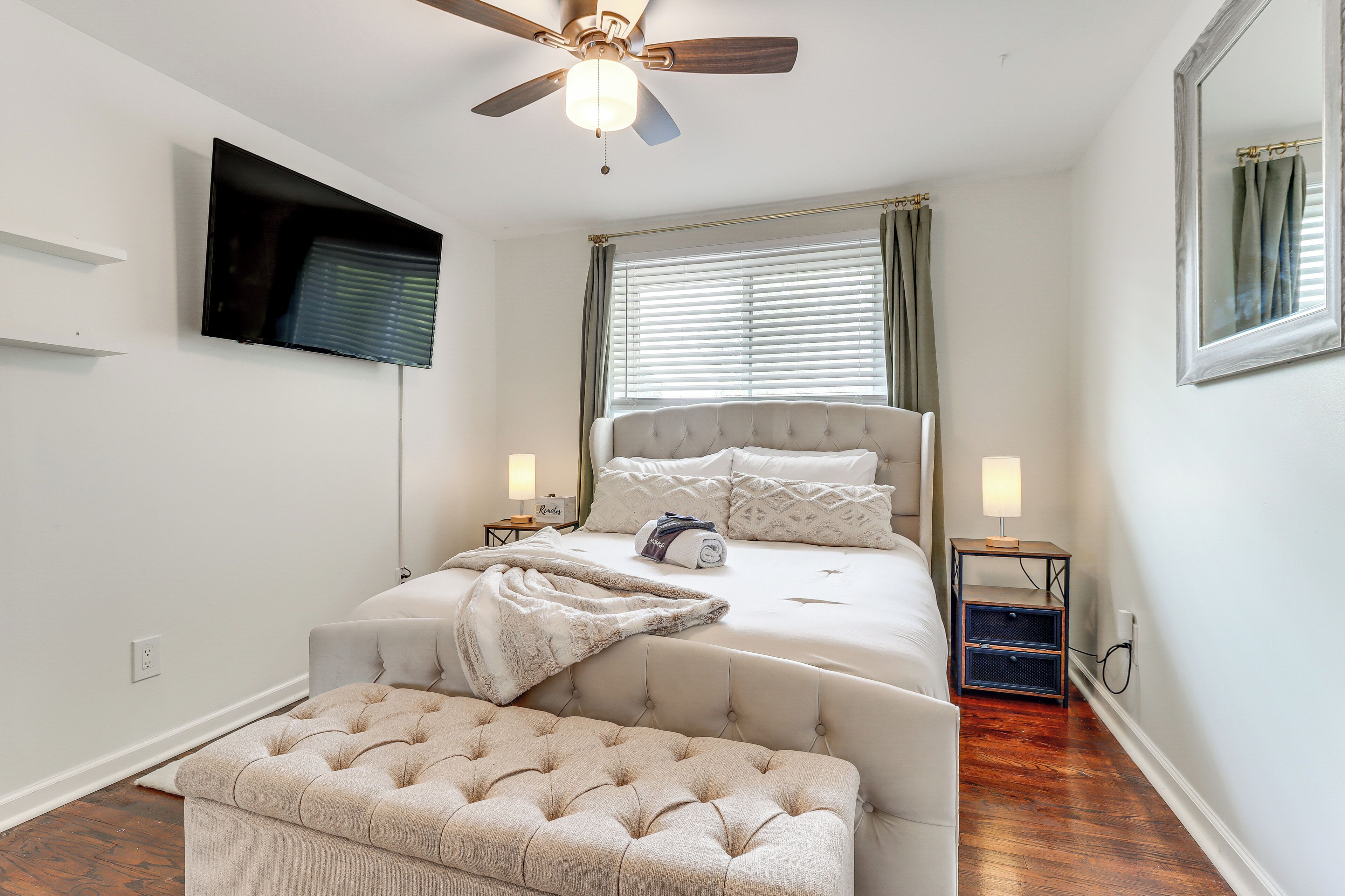 Une chambre avec un grand lit, un ventilateur de plafond, un téléviseur fixé au mur et une table de chevet avec une lampe.