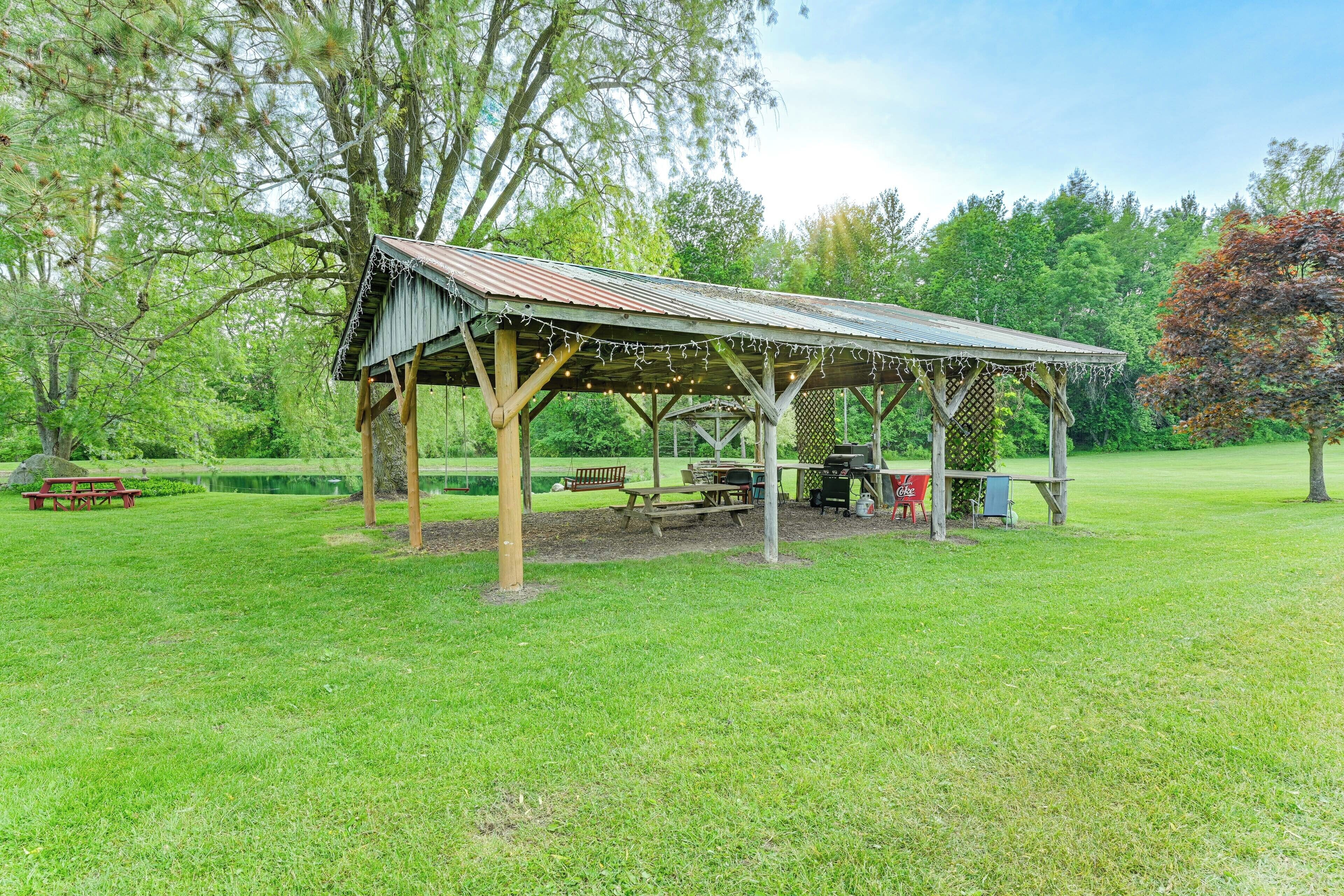 A covered picnic area with benches and picnic tables, surrounded by trees and a grassy field.