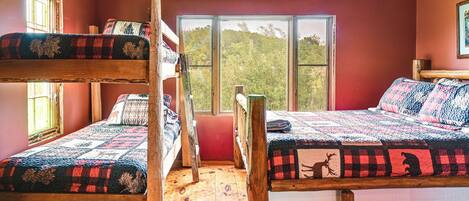 A room with two bunk beds, wooden flooring, and large windows offering a view of trees.