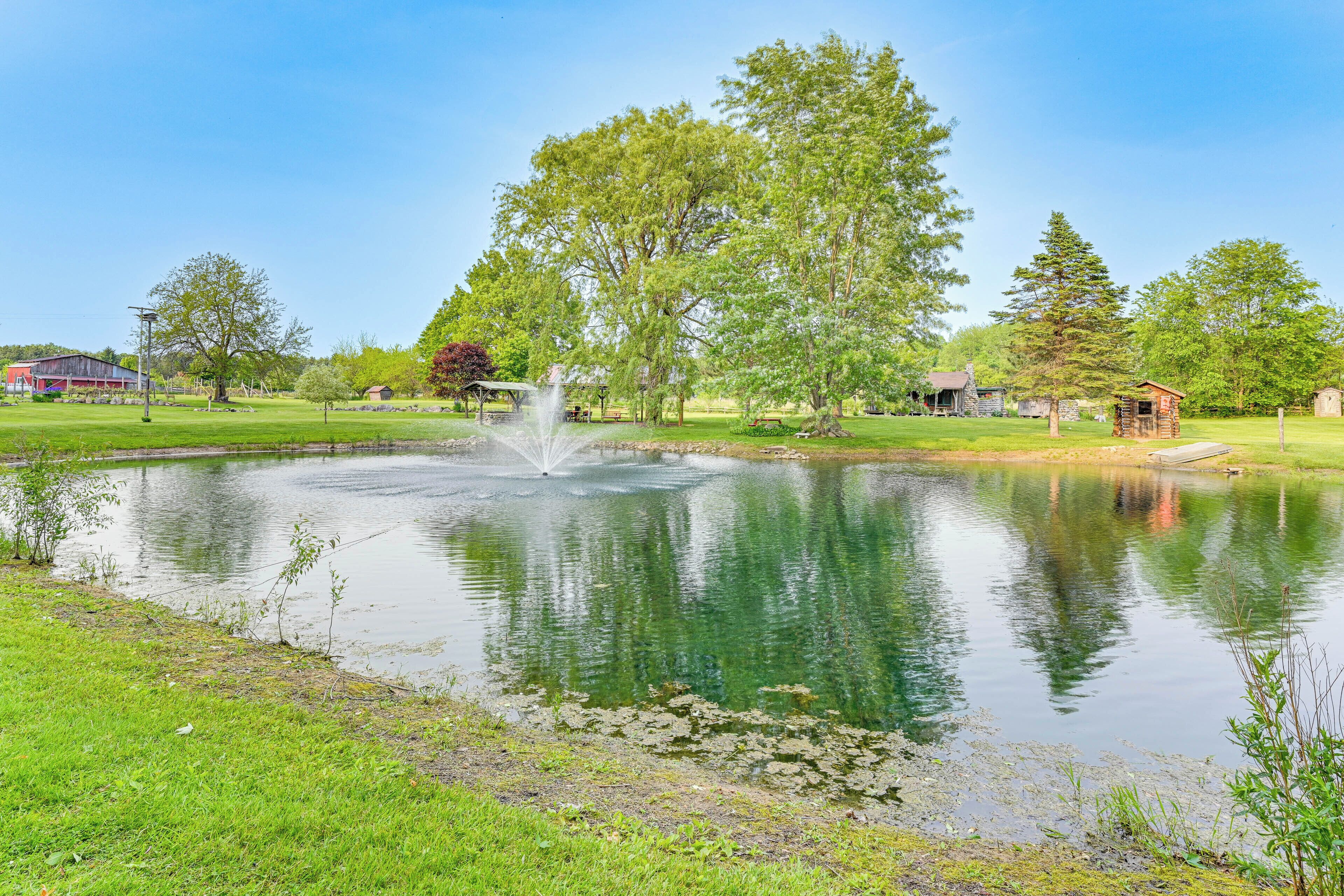 Un étang paisible avec une fontaine, entouré d’une végétation luxuriante et surplombé d’un ciel bleu clair.