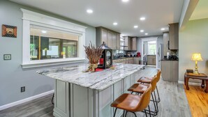 A kitchen with a marble island, wooden bar stools, and a view of the backyard through a large window.