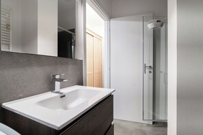 A modern bathroom with a white sink, dark cabinetry, a glass shower enclosure, and a wooden door. - Stylish 2-Bedroom Apartment by Lake Como (Como)