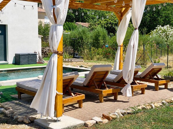 A poolside area with wooden lounge chairs and a pergola with white curtains. - Lorgues: 3-BR house, private pool, A/C & fiber Wi-Fi (Lorgues)