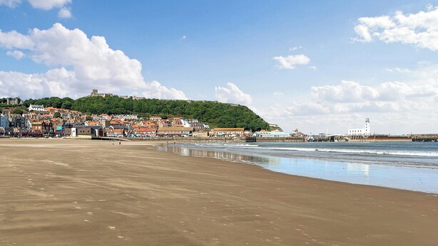A beach with a town in the background, a lighthouse, and a hill with buildings.