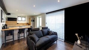 Ein modernes Wohnzimmer mit einer dunkelgrauen Couch, einem Holzesstisch mit Barhocker, einer Küchenzeile mit Mikrowelle und Kaffeemaschine sowie einem großen Fenster mit Jalousien.