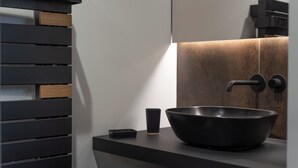 Ein modernes Badezimmer mit einem schwarzen Waschbecken, einer kleinen schwarzen Seifenschale und einem Holzdetail an der Wand.