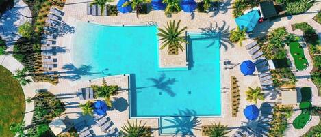 An aerial view of a resort with a central swimming pool, sun loungers, palm trees, and a golf course.