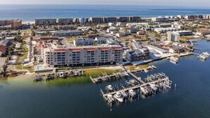 An aerial view of a coastal marina with a hotel featuring multiple floors and a red roof, surrounded by other buildings and a clear view of the sea.