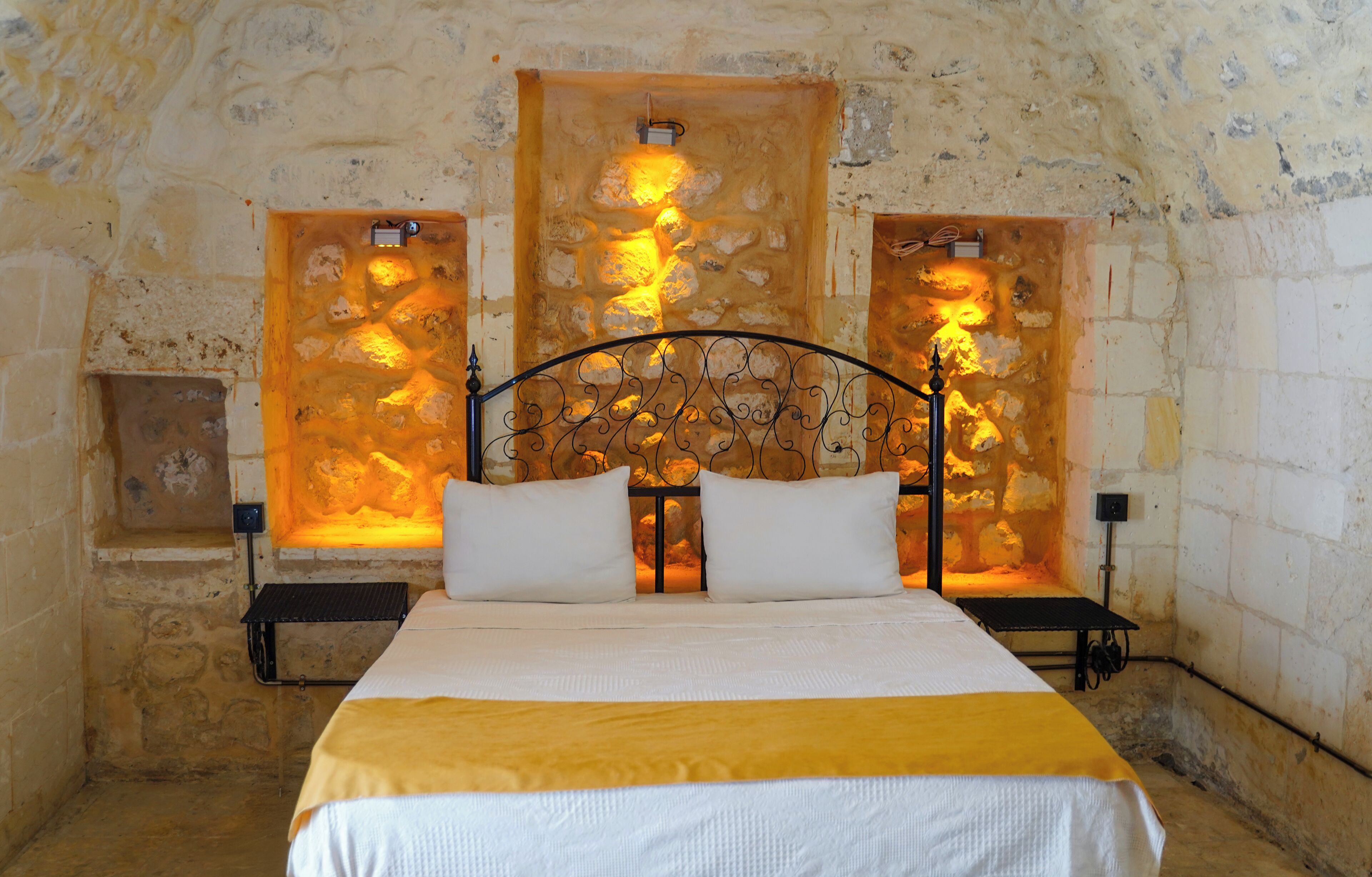 Una habitación con paredes de piedra, una cama, dos mesitas de noche y dos lámparas de pared.