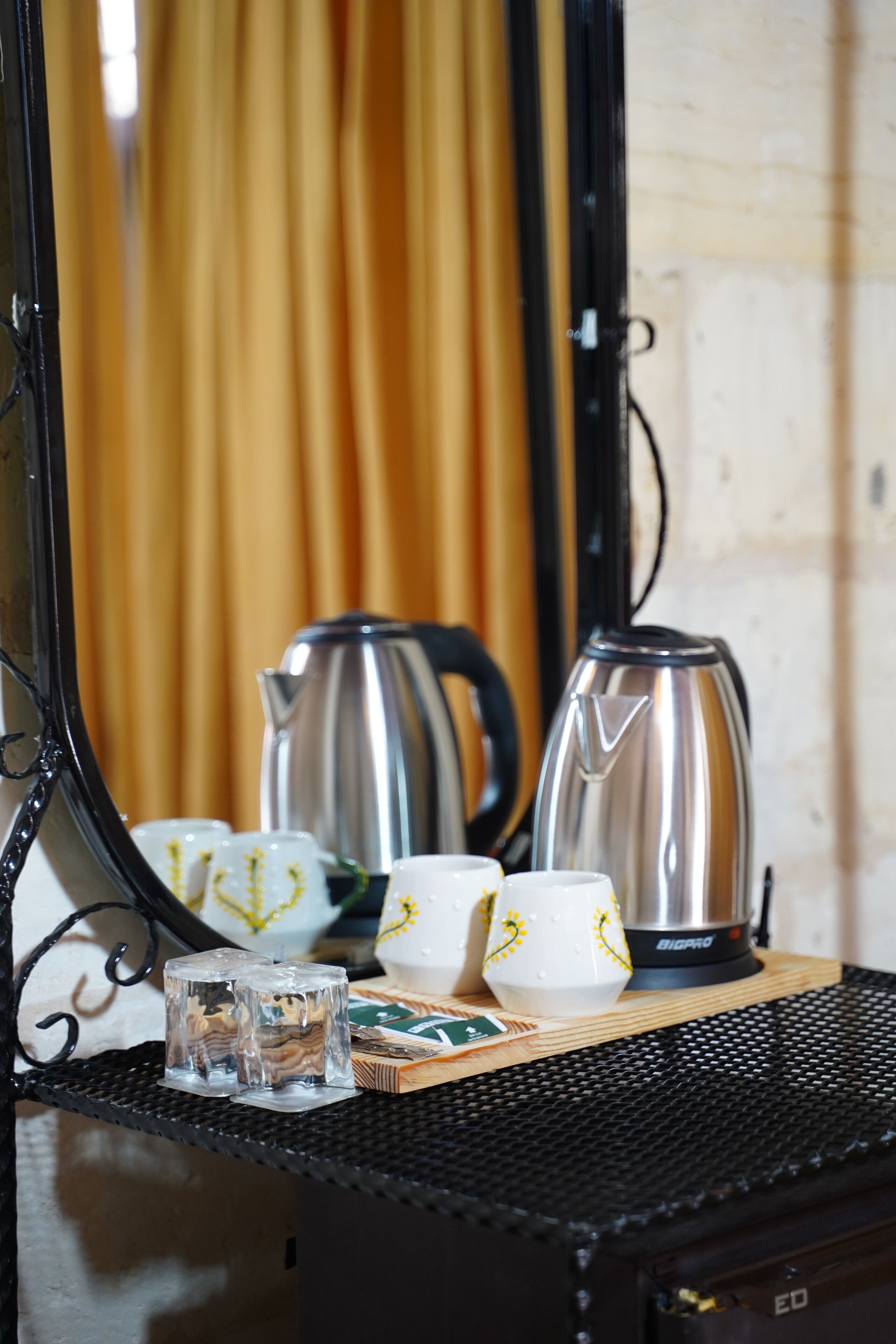 Une pièce avec un meuble en bois, une table en métal noir, deux bouilloires et un miroir qui reflète un rideau jaune.