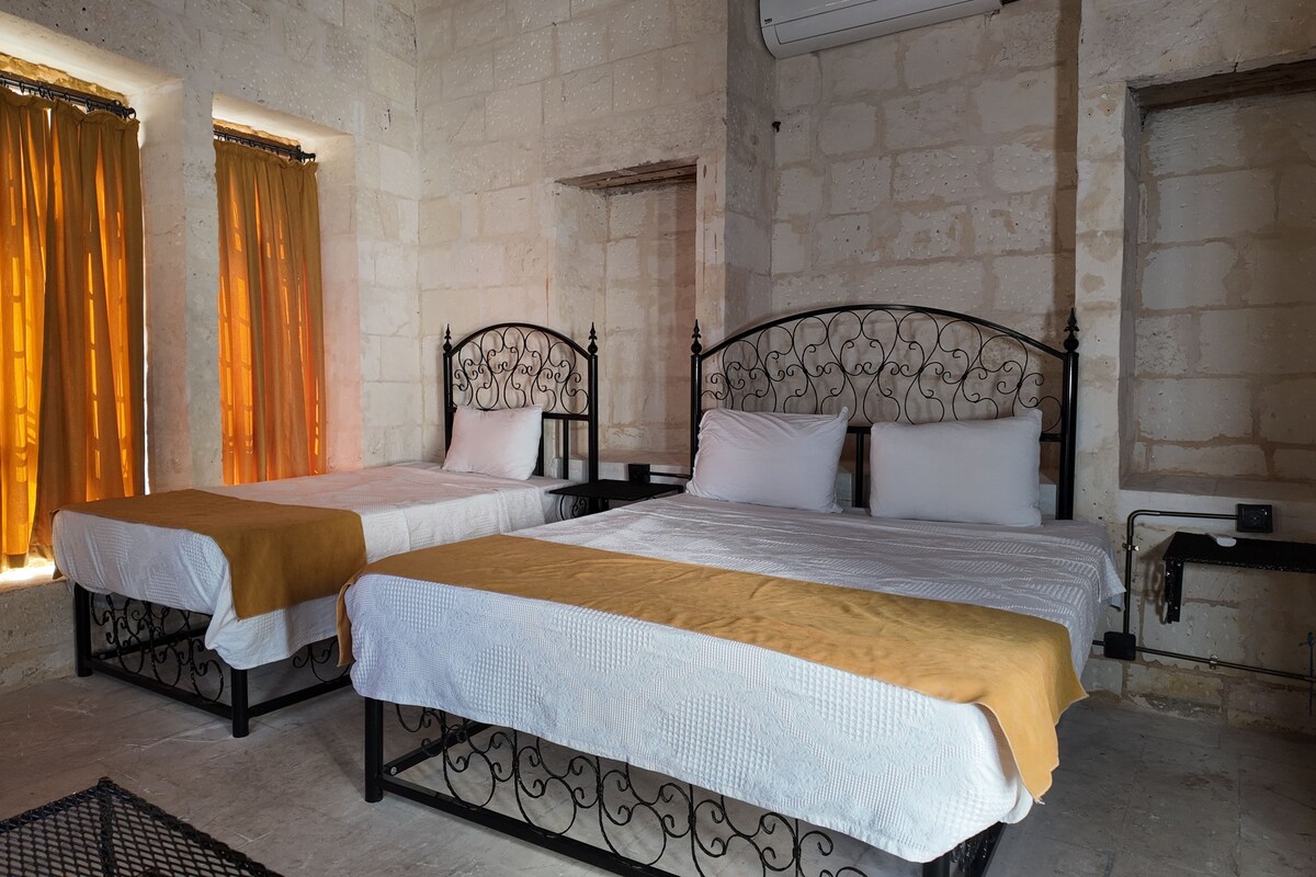 A room with two beds, a window with curtains, a wall-mounted air conditioner, and a metal table.