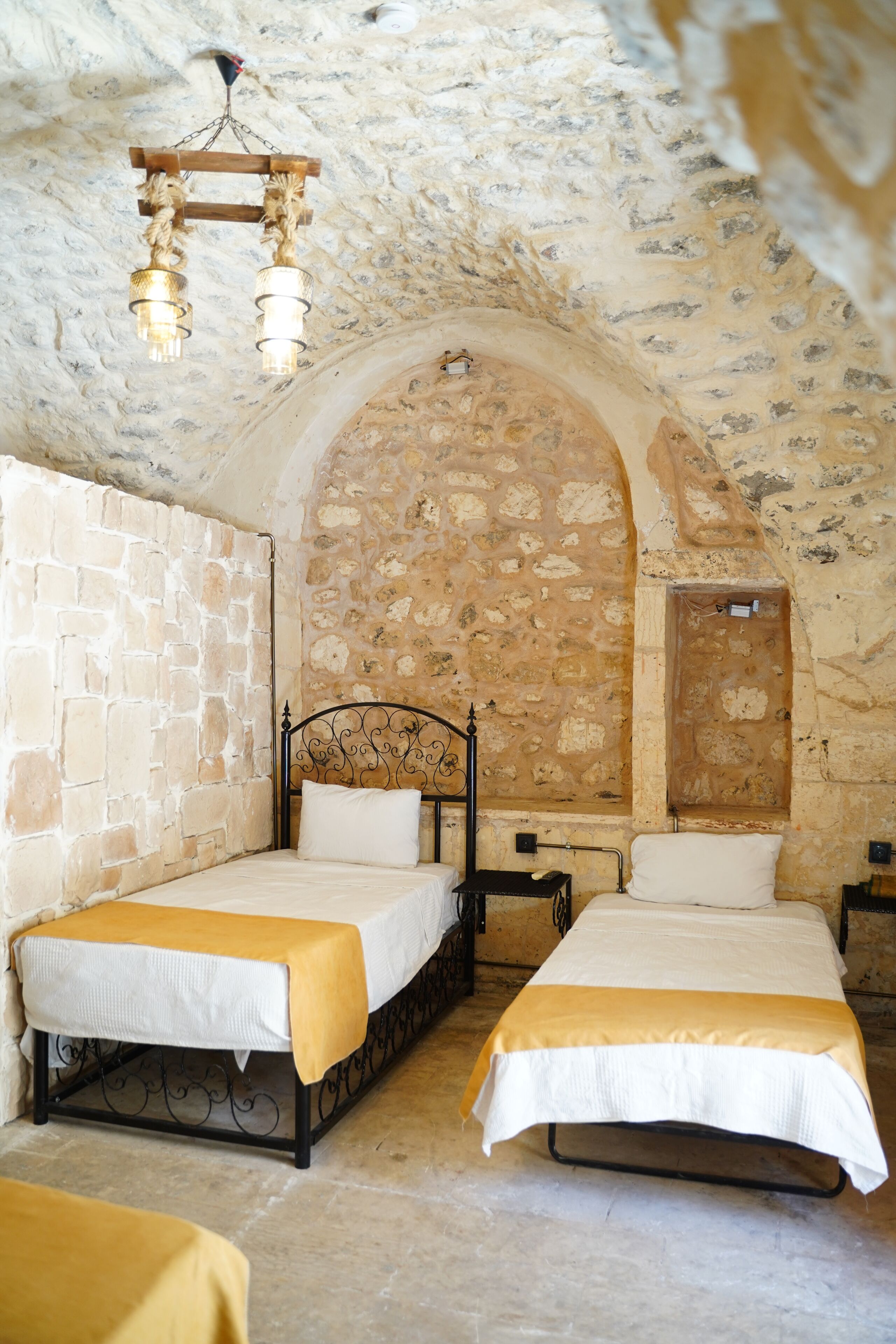 A stone-walled room with two beds, a hanging light fixture, and a wooden door.