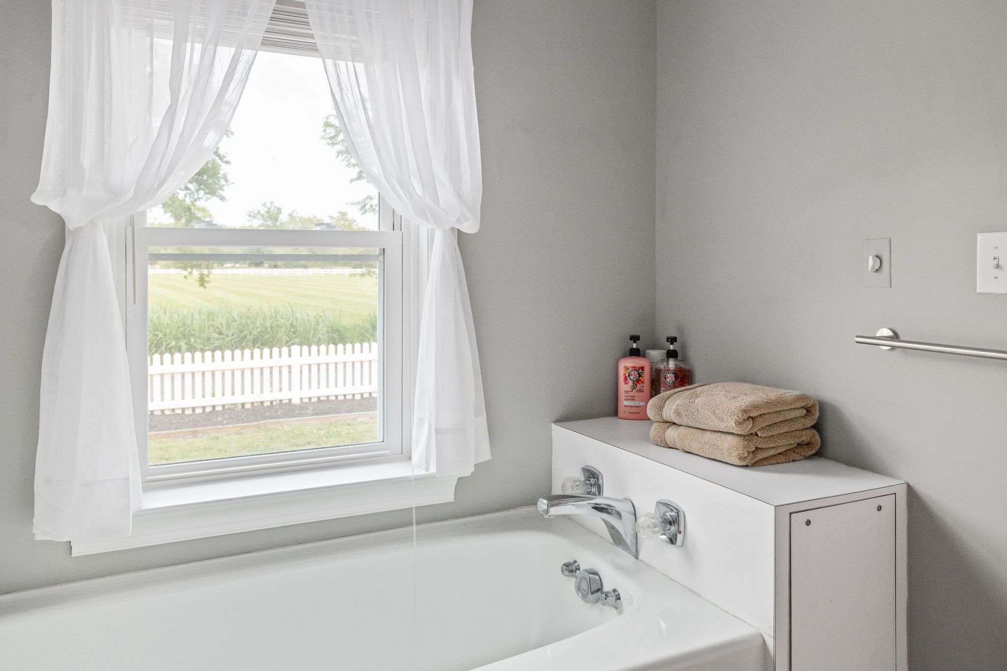 Un baño con una bañera blanca, una ventana con cortinas blancas y un perchero con toallas y artículos de aseo.