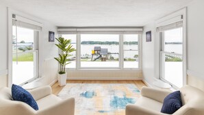 A room with a large window overlooking a body of water, a potted plant, and two sofas with cushions. - Bayfront Oasis with Heated Pool, Hot Tub & Playground! Family & Pet friendly! (Reedville)
