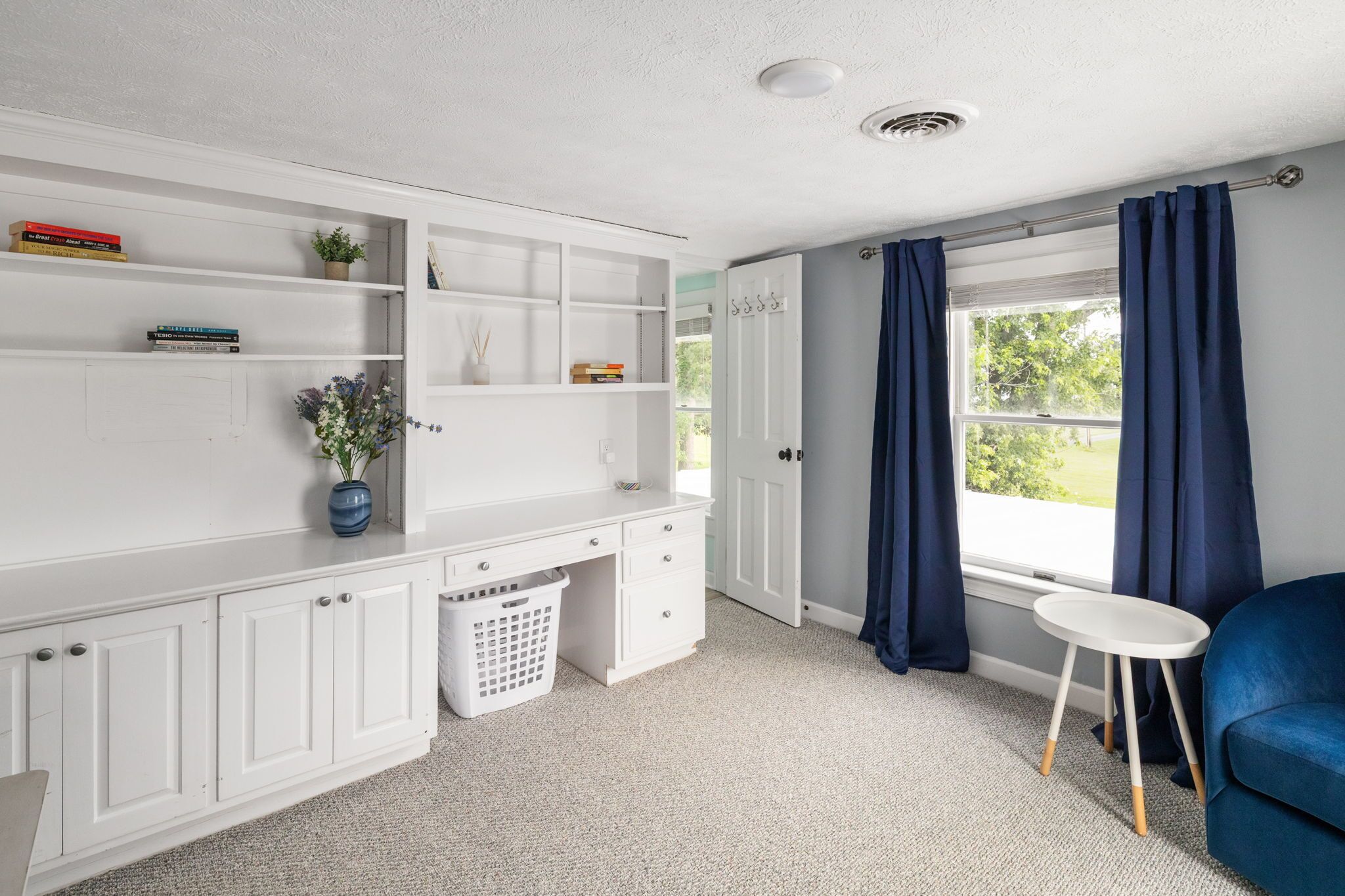 A room with a white desk, shelves, a small table, a blue chair, and a window with curtains.