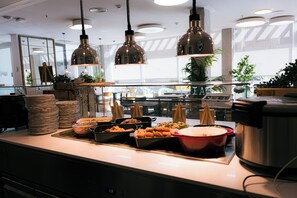 A buffet area with various food items, stacked plates, and hanging pendant lights. - Bravo Students Granada - Campus Accommodation - Campus Accommodation (Granada)