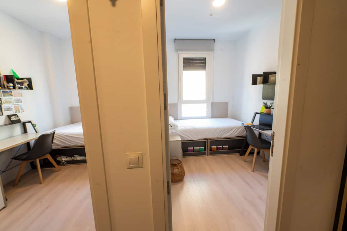 Una camera da letto con un letto, una scrivania e una sedia. Si vede una finestra con persiane, una porta e uno scaffale con diversi oggetti.