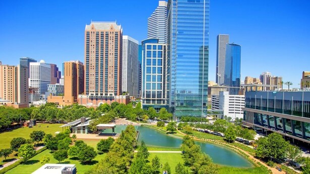 A cityscape with modern high-rise buildings, a central park with a pond, and a clear blue sky.
