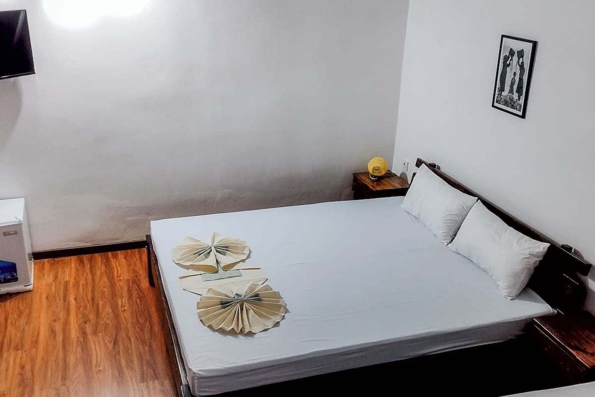 Quarto com cama, televisão na parede, mesinha de cabeceira com abajur e quadro pendurado na parede.