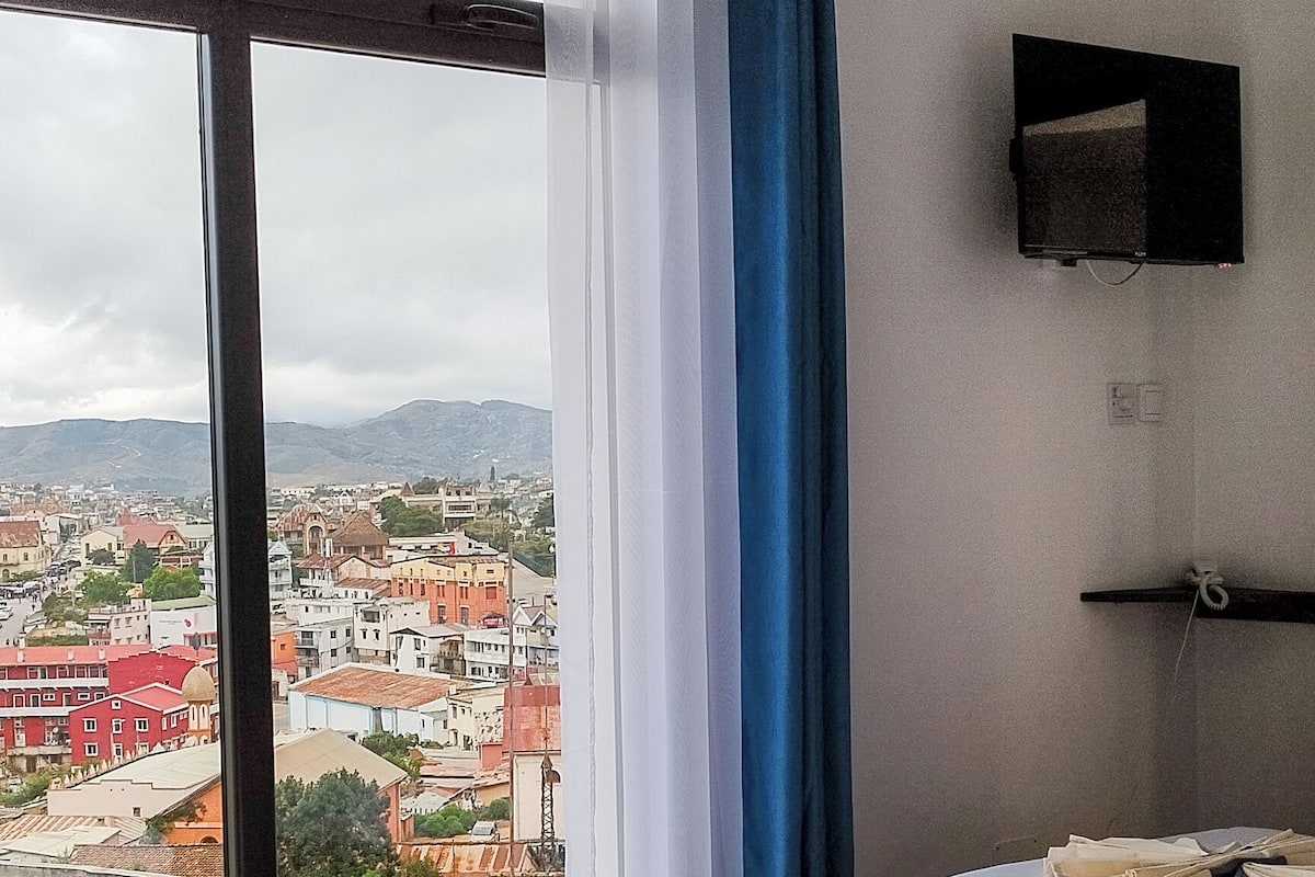 Quarto de hotel com duas camas, um ventilador de teto, televisão na parede e uma janela grande com vista para a cidade.