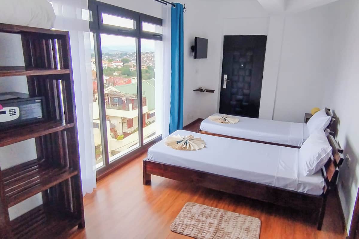 Quarto com duas camas, uma estante de madeira com um telefone e vista da cidade por grandes janelas.