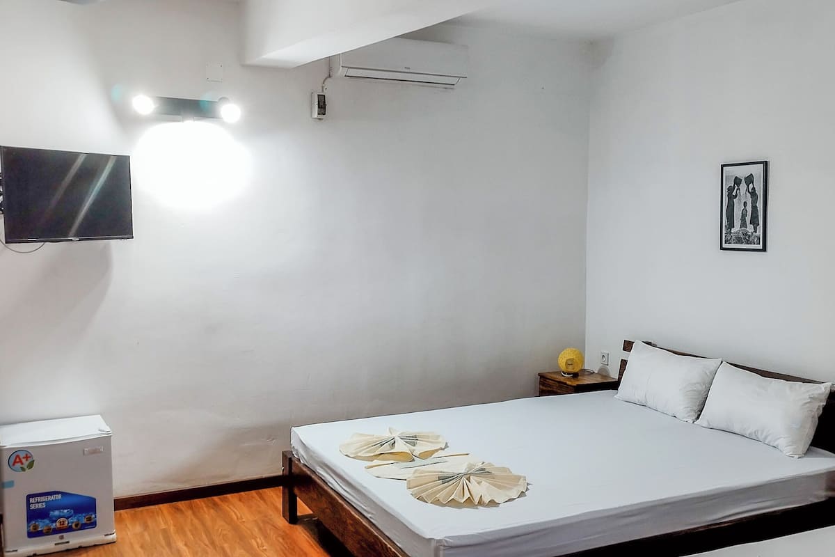 Quarto de hotel com cama, mesa de cabeceira, televisão fixada na parede, ar condicionado e quadro decorativo na parede.