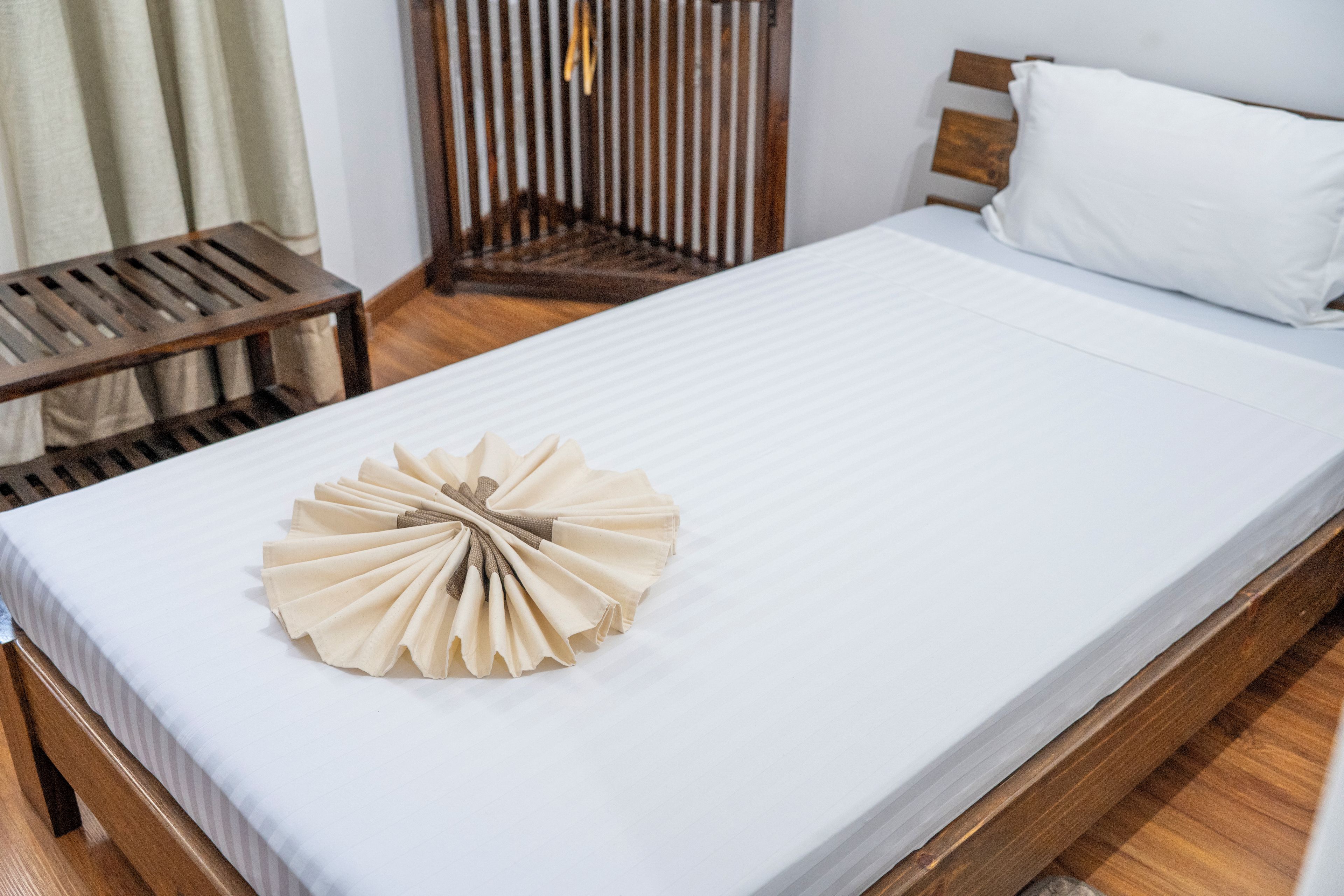 Una cama bien tendida con una toalla doblada en el centro, un cabecero de madera y una mesita de noche de madera con una lámpara.