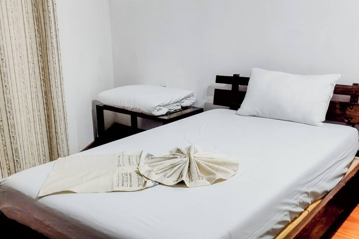Quarto de hotel com cama bem arrumada, mesa de cabeceira com abajur e janela com cortinas.