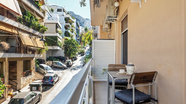 Une rue étroite bordée de voitures garées, un balcon avec une table et des chaises, et des immeubles dotés de balcons et de plantes.