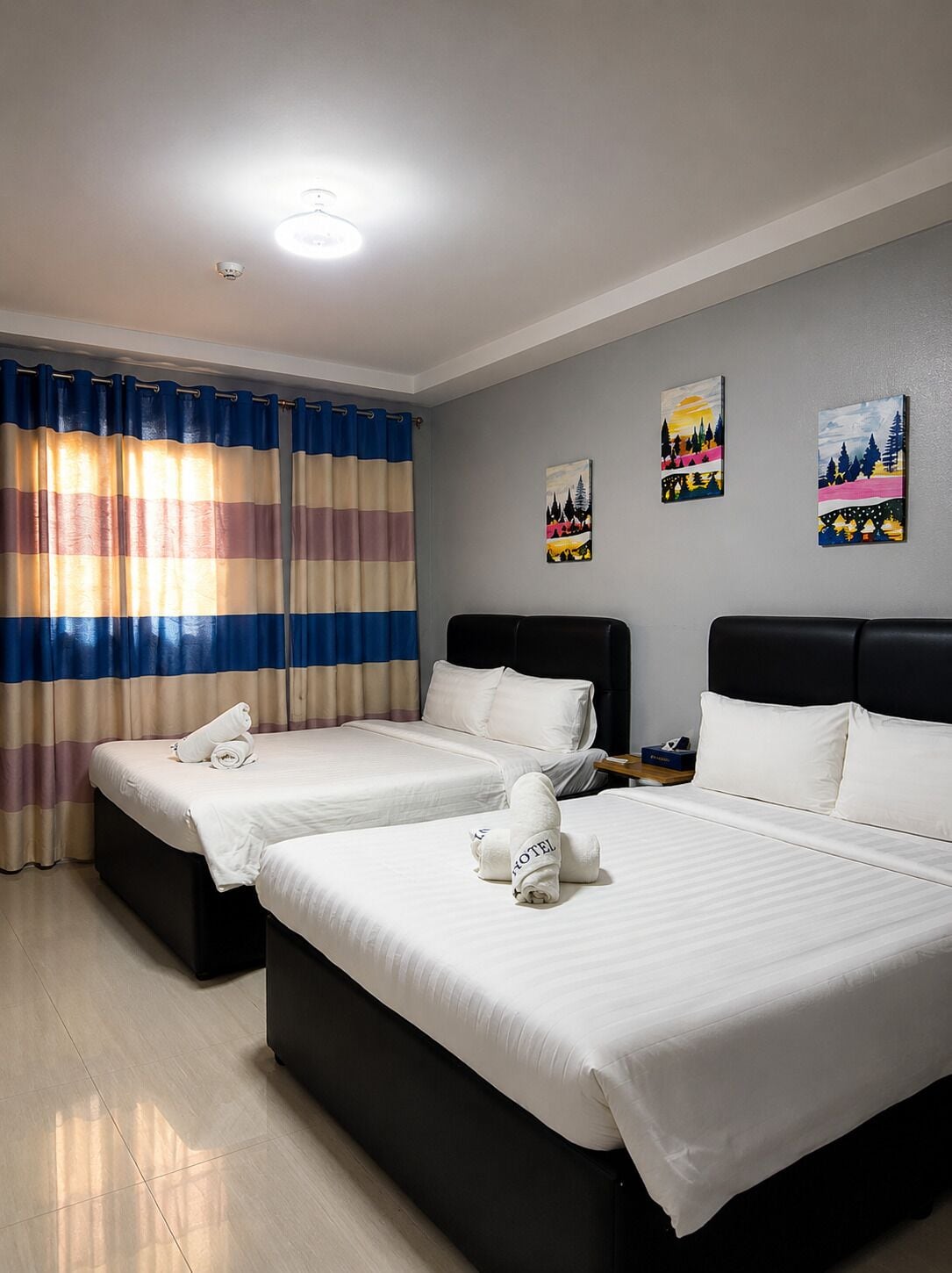 A hotel room with two beds, a large window with curtains, a ceiling light, and three paintings on the wall.