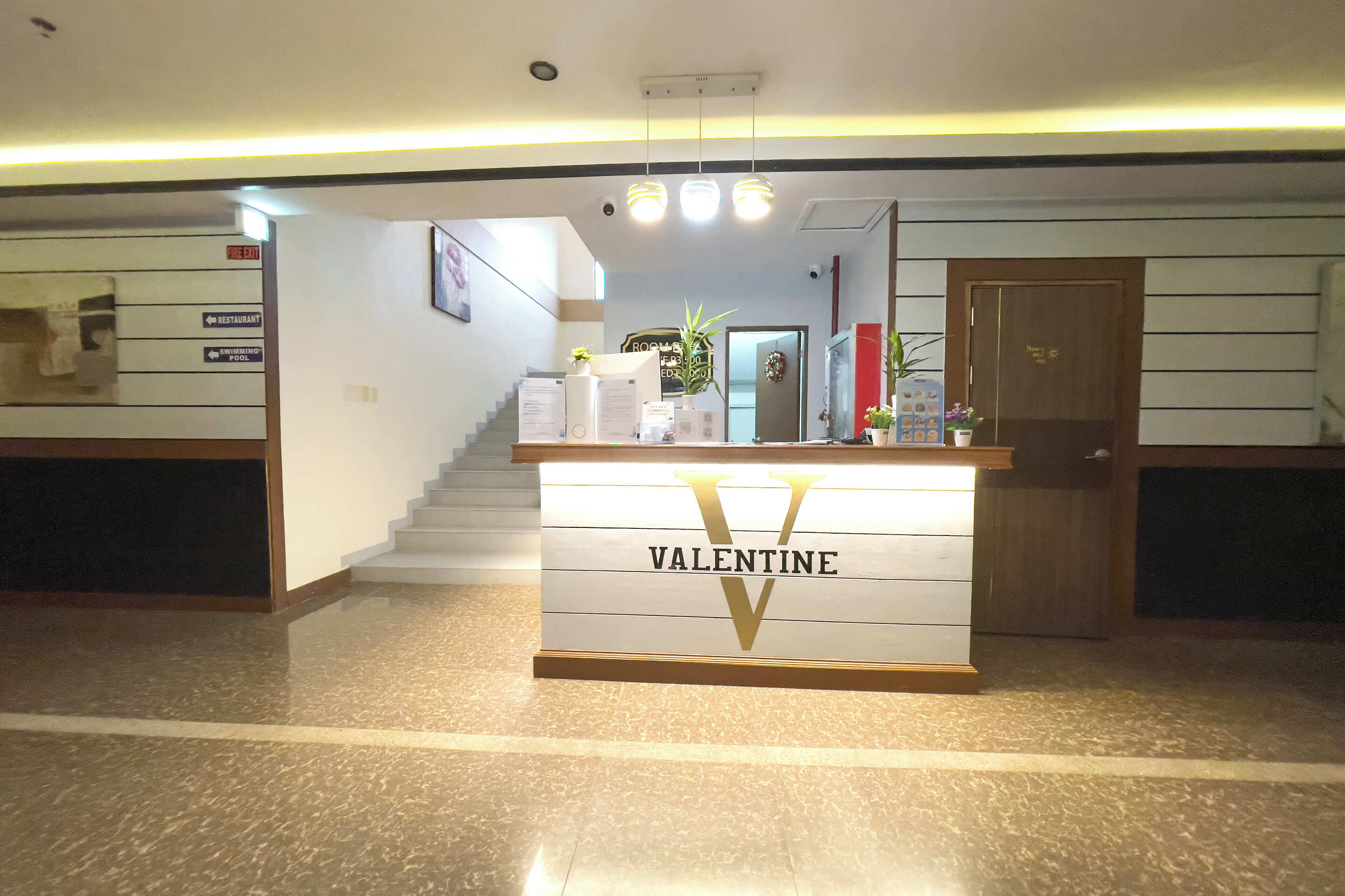 A reception desk with a sign reading "Valentine" in a hotel lobby with a staircase, a sign for a restaurant, and a pool.