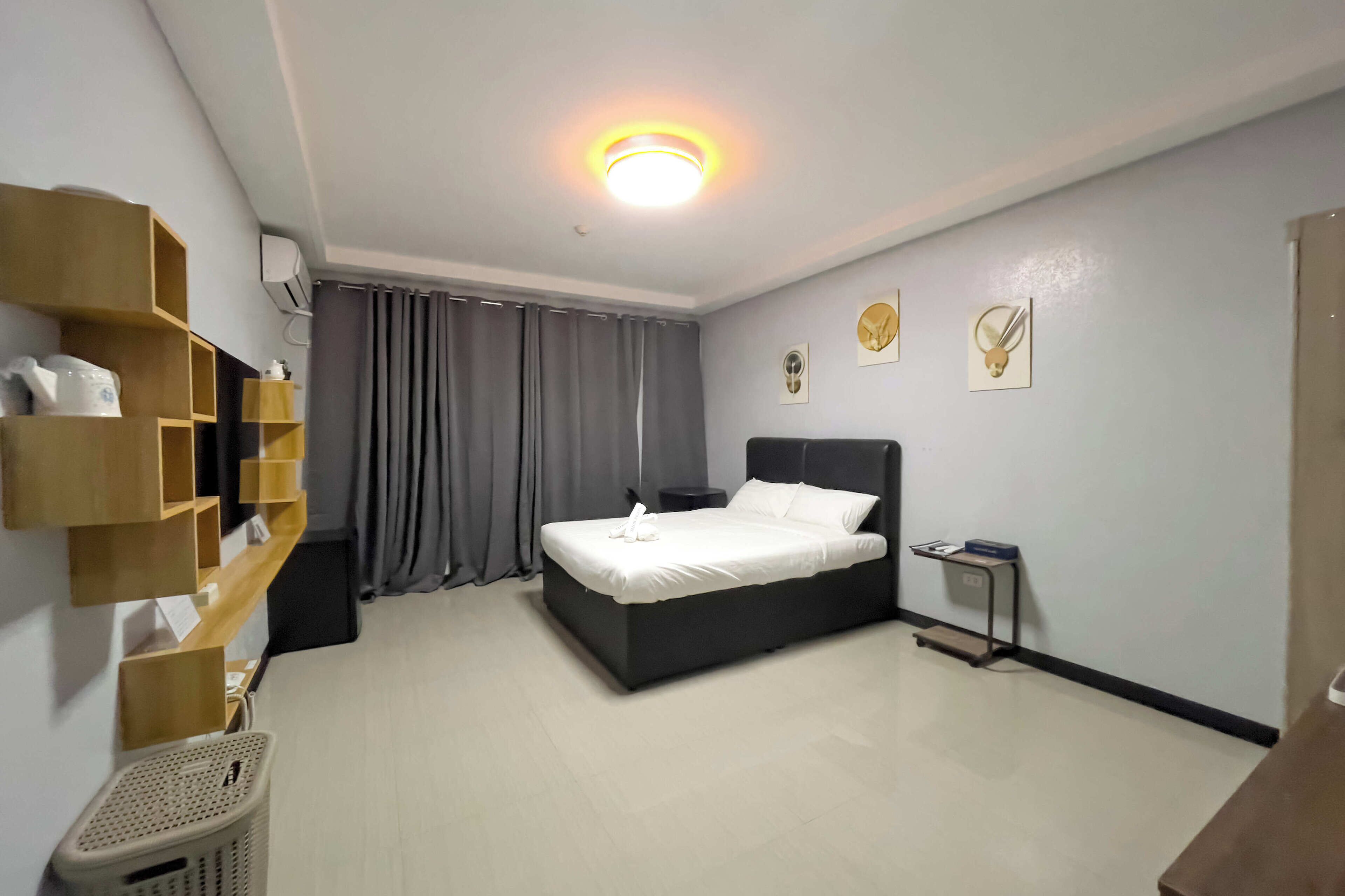 Habitación de hotel con cama, una mesita de noche, estante en la pared, cesto de ropa y una lámpara en el techo.
