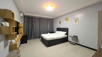 Habitación de hotel con cama, una mesita de noche, estante en la pared, cesto de ropa y una lámpara en el techo.