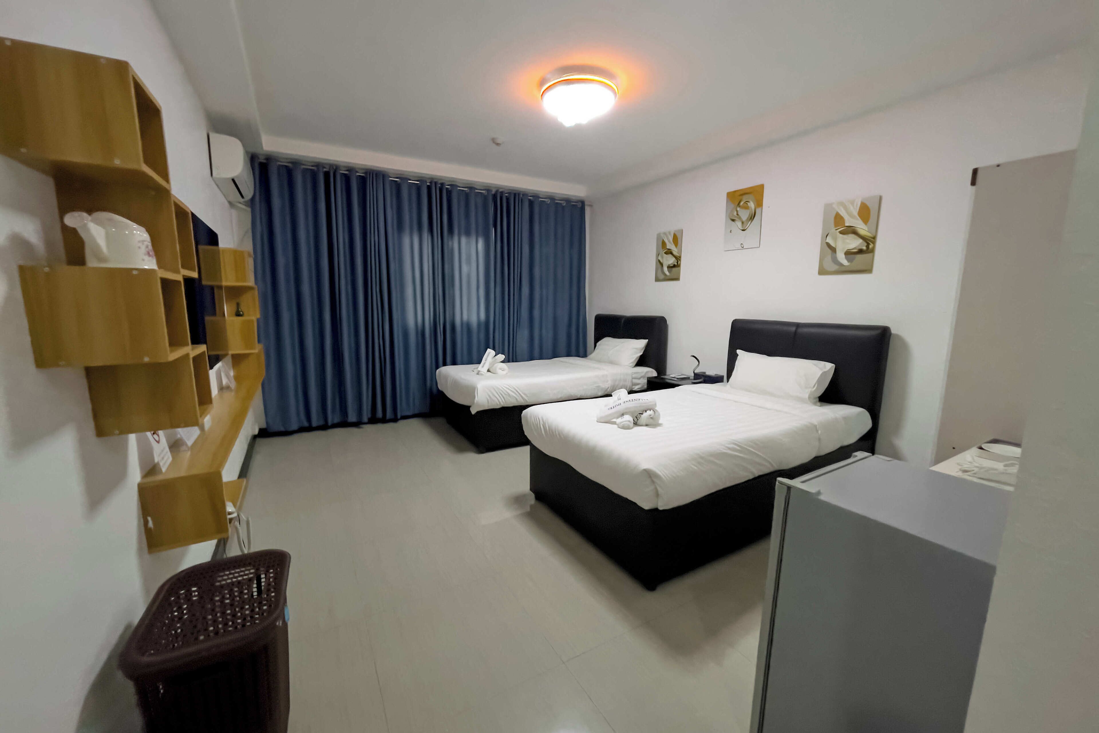 Habitación de hotel con dos camas, un escritorio, una silla, un cesto de basura y un estante en la pared con objetos.