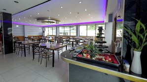 A modern dining area with tables and chairs, a buffet counter with fruits and vegetables, and large windows allowing natural light. - Valentine Hotel Angeles City managed by Cocotel (Angeles City)