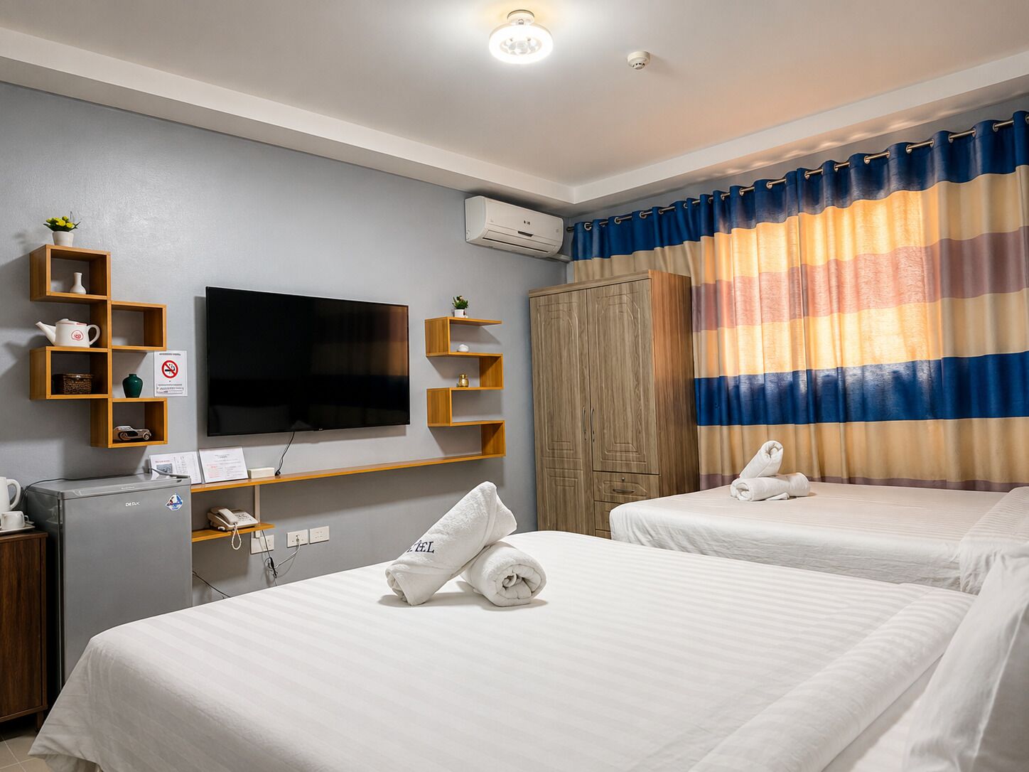 A hotel room with a flat-screen TV, wooden shelves, a wardrobe, and a striped curtain.