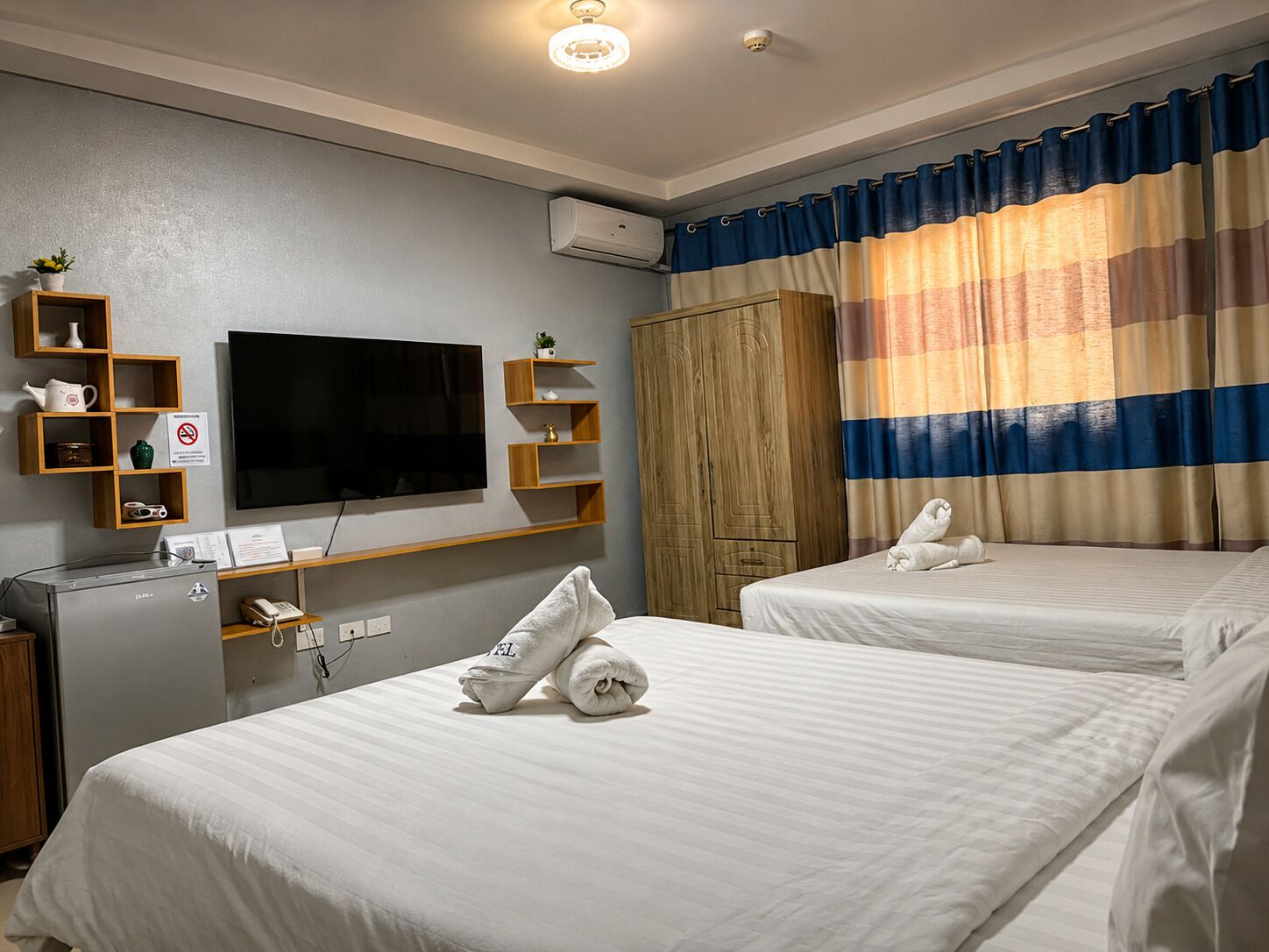 Habitación de hotel con dos camas, televisor, armario y una pequeña estantería con artículos de aseo.