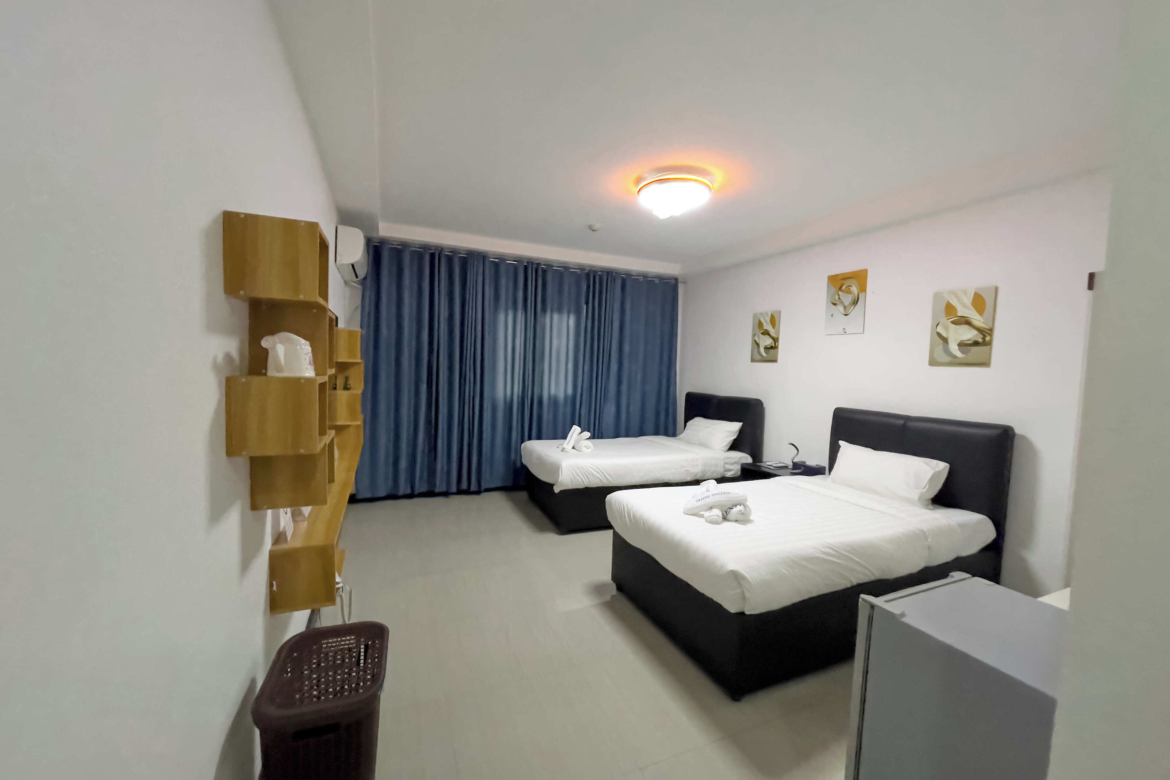 A hotel room with two beds, a desk, a chair, a trash bin, and a wall-mounted shelf.