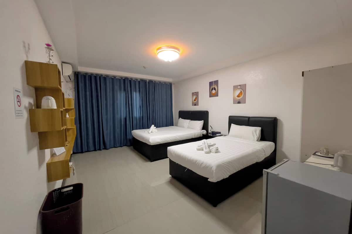 A hotel room with two beds, a desk, a trash bin, and a ceiling light.