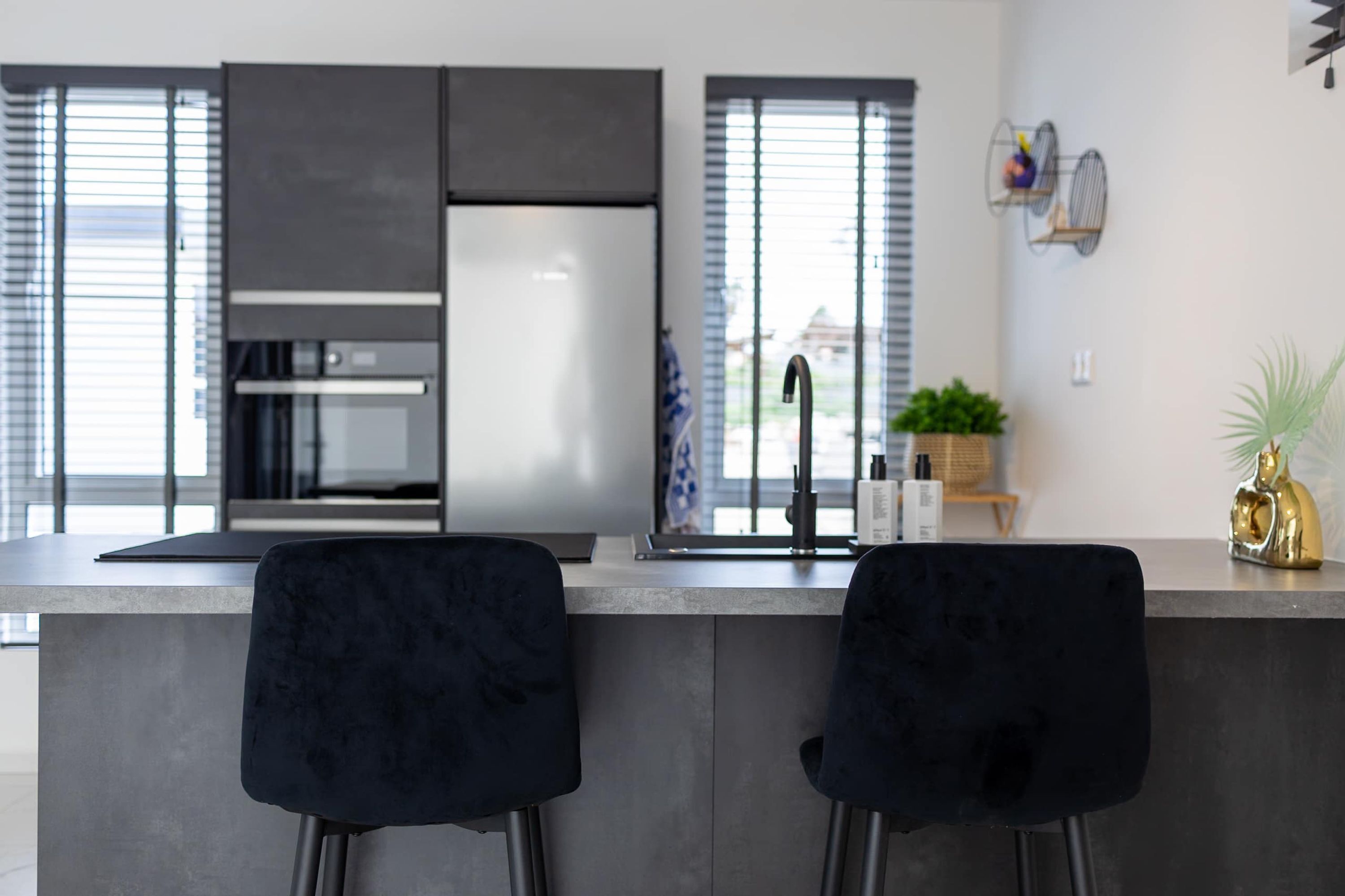 A modern kitchen with a central island, two black chairs, a stainless steel refrigerator, and a window with blinds.