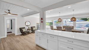 Una cocina amplia con armarios blancos, una isla grande e iluminación colgante. Hay un comedor con mobiliario de mimbre y vistas al exterior.