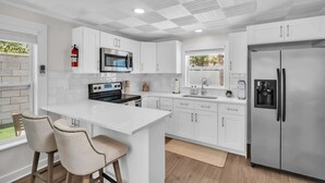 A modern kitchen with white cabinetry, a stainless steel refrigerator, a microwave, and a stove. There are two bar stools and a window with a view of greenery.
