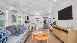 A modern kitchen with white cabinets, a wooden dining table, and a flat-screen TV mounted on the wall.