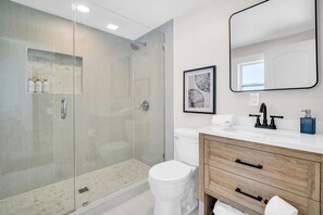A modern bathroom with a glass-enclosed shower, a white toilet, a wooden vanity with a sink, and a framed picture on the wall. - New Sweet Beach Nook Hot Tub Ocean Vibes (Indian Rocks Beach)