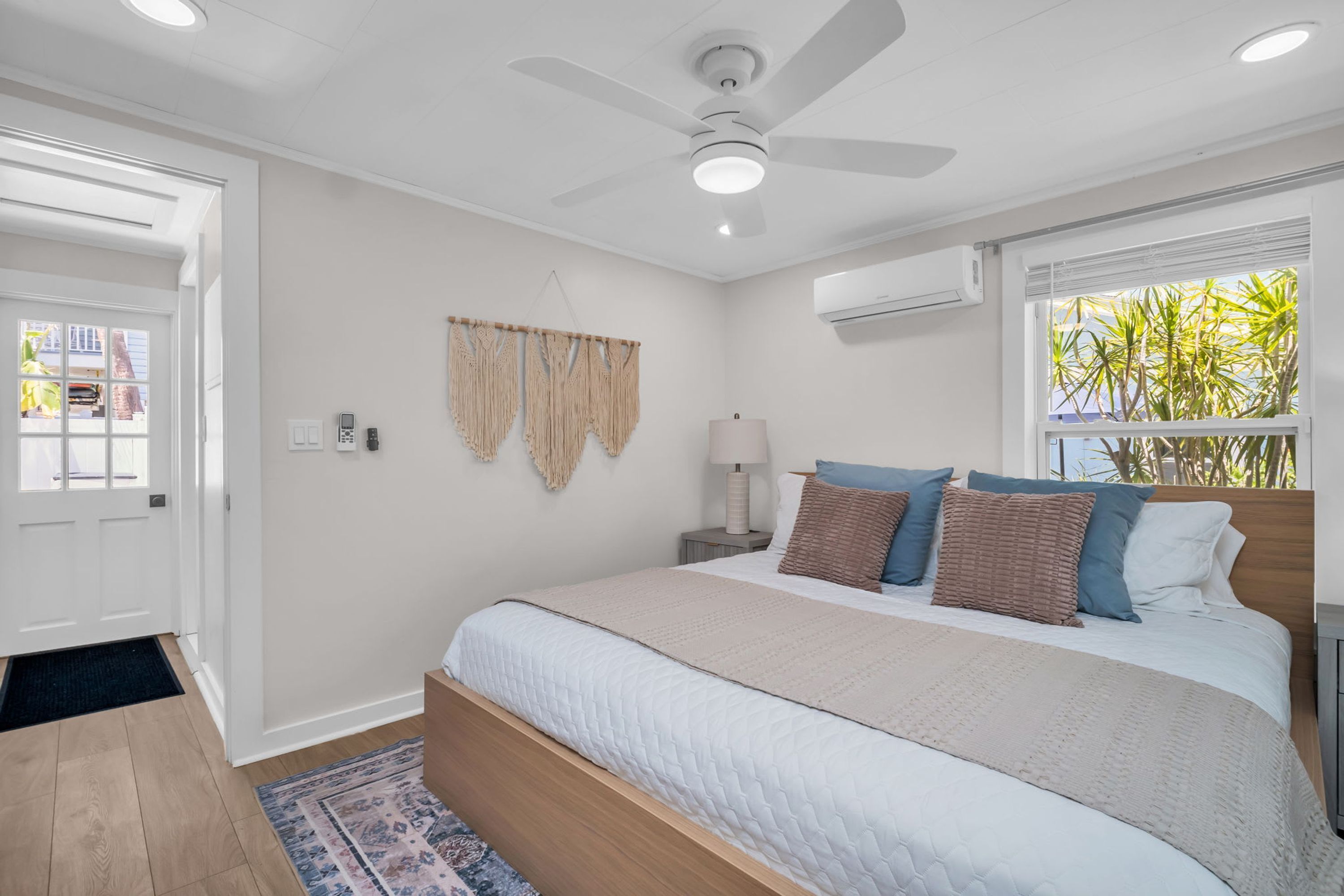 A bedroom with a bed, a ceiling fan, an air conditioning unit, a window with plants, and a door.