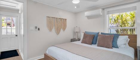 A bedroom with a bed, a ceiling fan, an air conditioning unit, a window with plants, and a door.